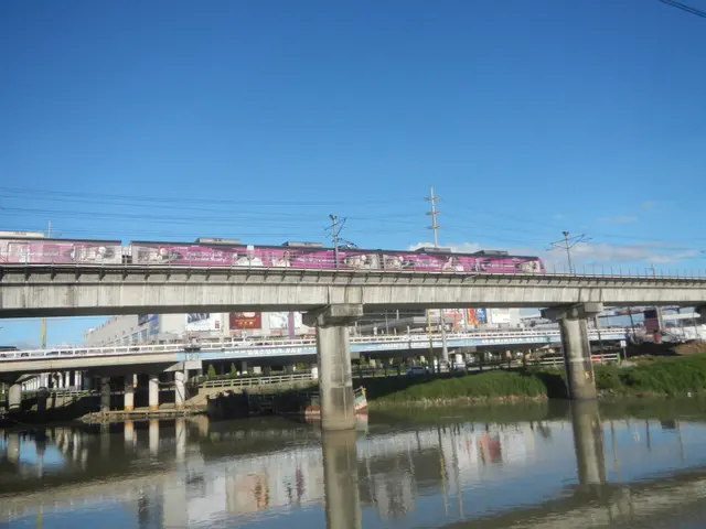 The image shows a train traveling over a bridge over a body of water, with buildings, poles, wires,...