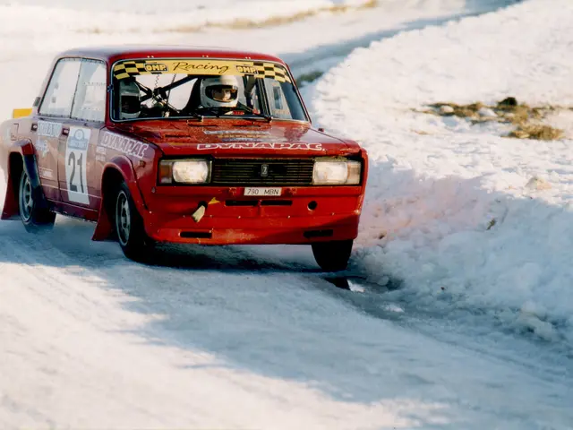 The image shows a red car driving down a snowy road, with two people inside wearing helmets. The...