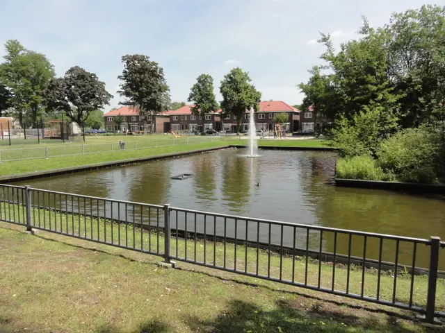 The image shows a pond with a fountain in the middle of it surrounded by a metal fence, grass,...