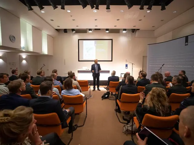 The image shows a group of people sitting in chairs in front of a projector screen, with a man...