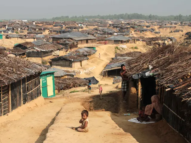 The image shows a group of people sitting on top of a dirt road in a slum area, surrounded by huts,...