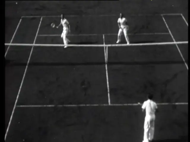 The image shows two men playing tennis on a black and white court. They are both holding rackets in...