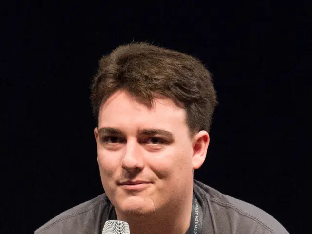 The image shows a man sitting on a chair, holding a microphone in his hand. He is wearing a black...