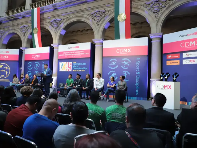 The image shows a group of people sitting on chairs in front of a stage, with a man standing beside...