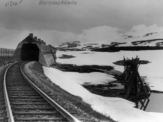 The image shows a black and white photo of a train track winding through a snowy landscape, with a...