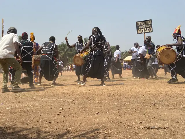 The image shows a group of people playing drums in a field surrounded by trees and a clear blue...
