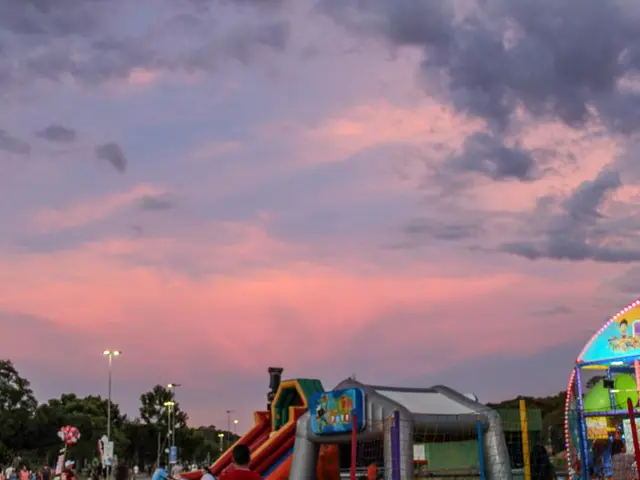 The image shows a crowd of people walking around a carnival at sunset, with a slide, amusement...