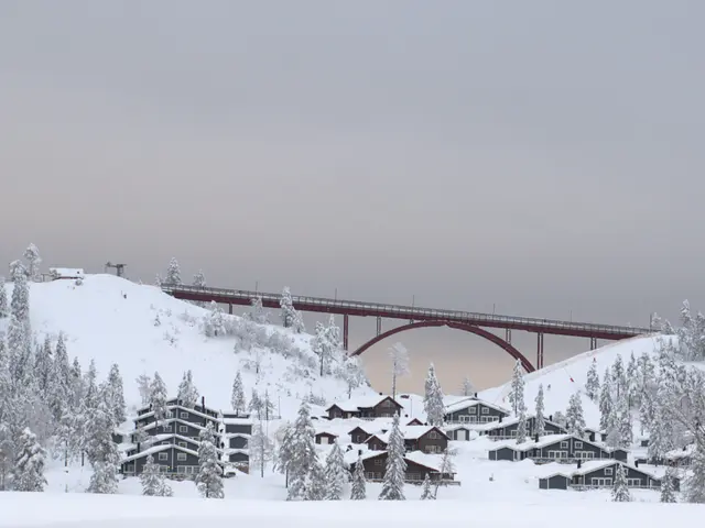 The image shows a picturesque winter scene of a ski resort with a bridge over a snowy hill. The...