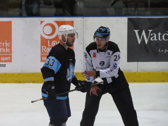 The image shows two men playing a game of ice hockey on an ice rink, both wearing helmets and...