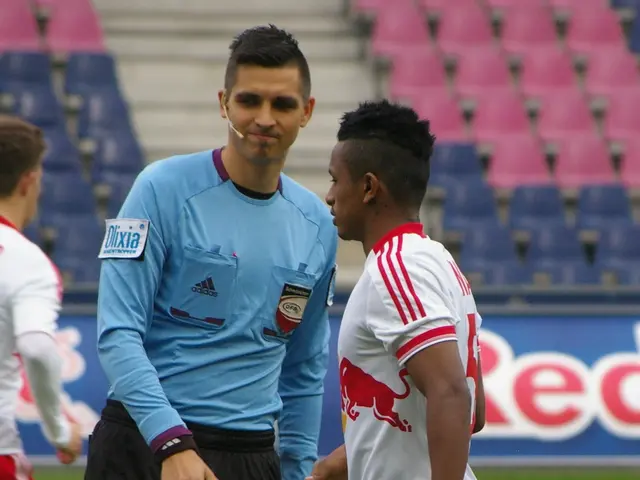 The image shows a referee talking to a soccer player on the field, surrounded by a group of people...