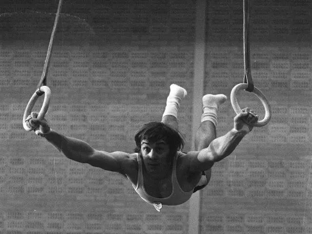 The image shows a man performing gymnastics on the rings in a gym, with a wall in the background...