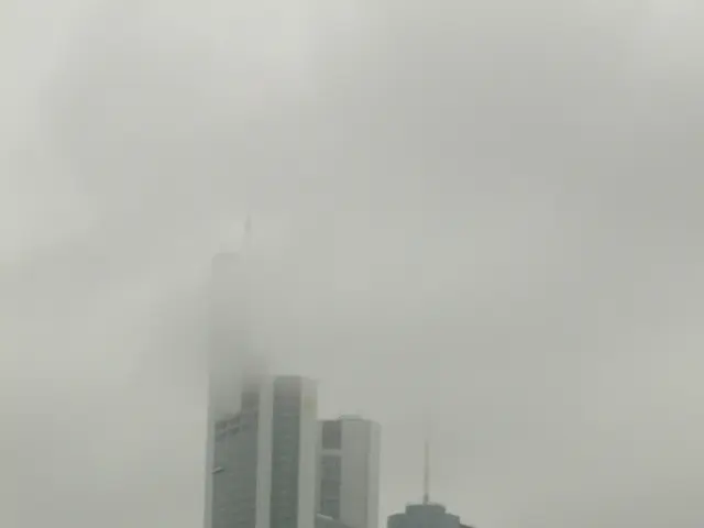 The image shows a foggy cityscape with tall skyscrapers in the background, surrounded by trees and...