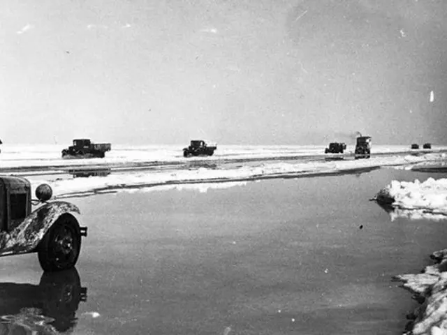 The image shows a black and white photo of a group of trucks driving down a frozen road, with snow...
