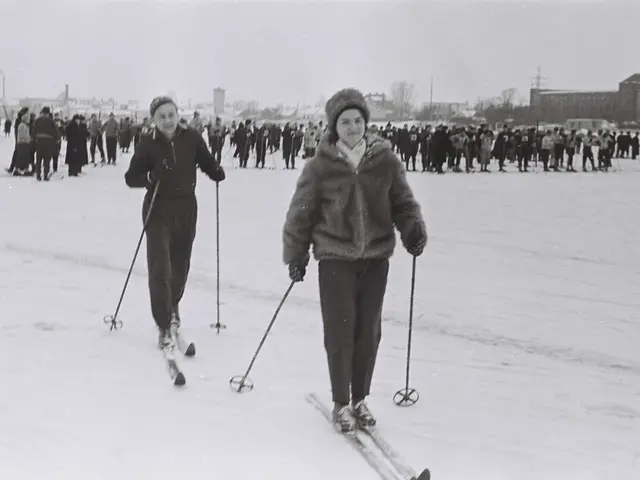 The image shows two people cross country skiing in the snow, wearing jackets, caps, gloves, and...