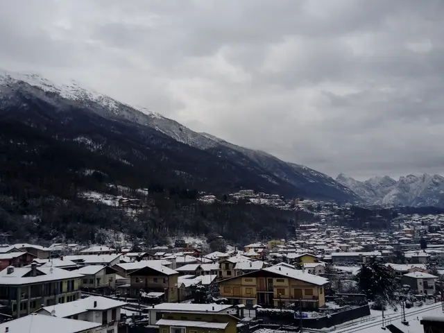 The image shows a small town with snow-covered mountains in the background. At the bottom of the...