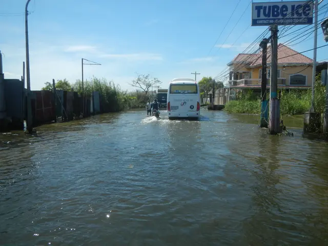 The image shows a bus driving through a flooded street with people on it. On either side of the...