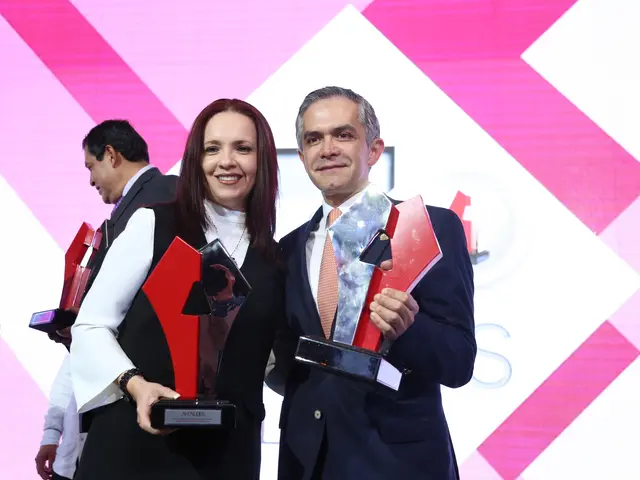 The image shows a man and woman standing next to each other on a stage, both of them smiling and...