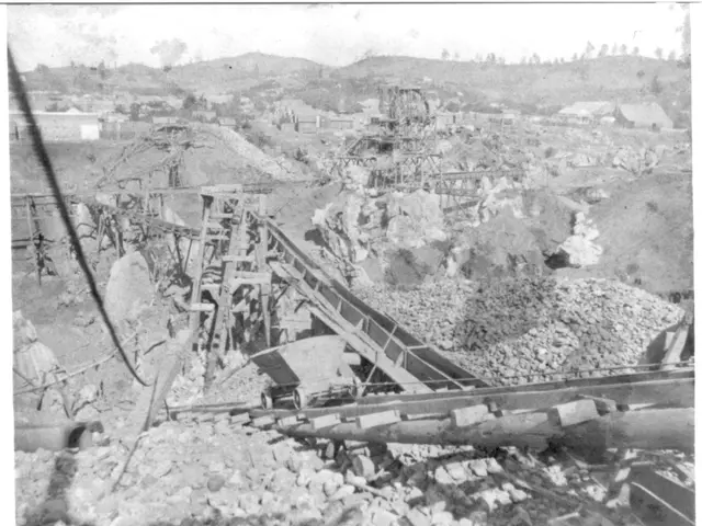 The image shows a black and white photo of a coal mine in Columbia, with a few buildings, trees,...