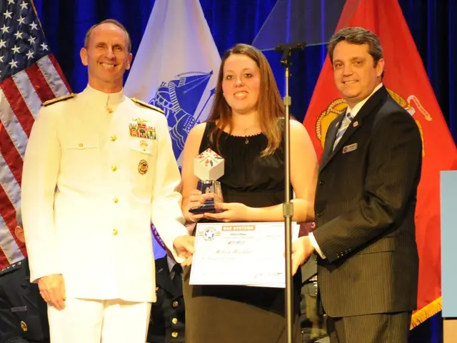 The image shows two men and a woman standing next to each other on a stage. The woman is wearing a...