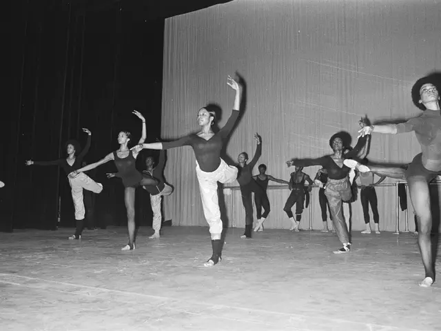The image shows a group of dancers on a stage with their arms outstretched, wearing black and white...
