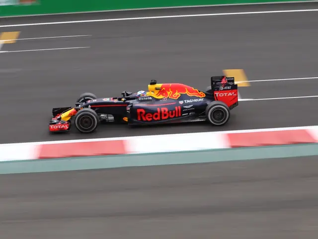 The image shows a red bull racing car driving on a race track, with the car in focus and the...