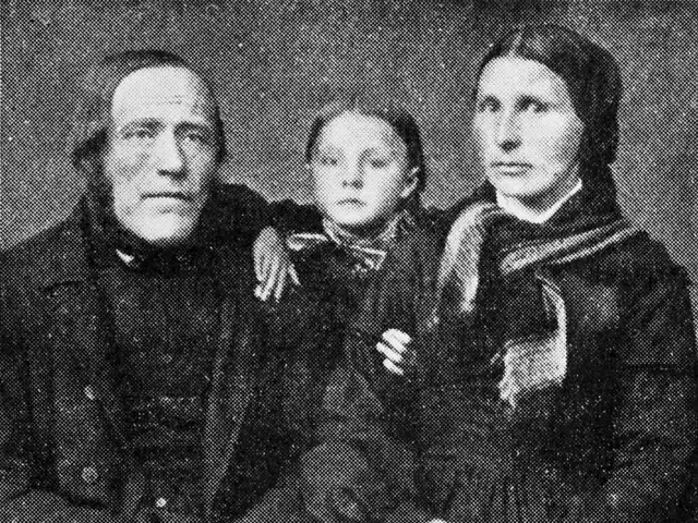 The image shows an old black and white photo of a family of three - a man, woman and a child -...