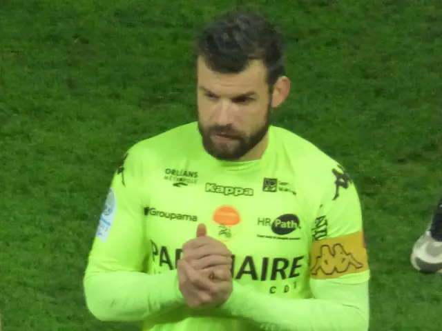 The image shows a man in a yellow goalkeeper's uniform standing on a soccer field, clapping his...