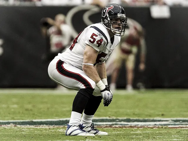 The image shows a football player kneeling on the field during a game, wearing a sports uniform,...
