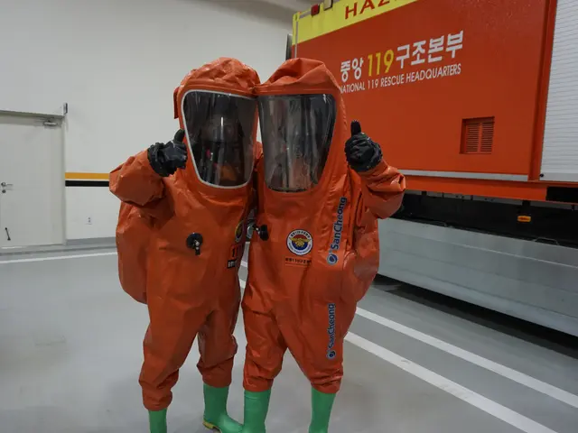 The image shows two people in orange hazmat suits standing next to each other in front of a truck,...
