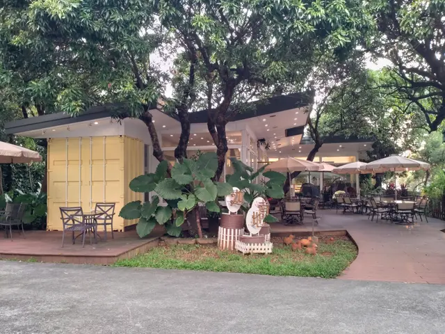 The image shows a restaurant with tables and chairs under umbrellas in the middle of a park,...