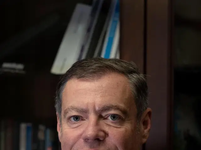 The image shows a man in a suit standing in front of a book shelf filled with books. He is...