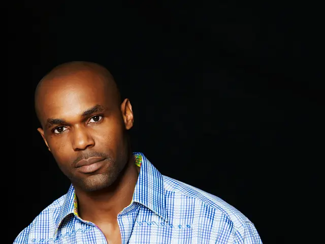 The image shows a man in a blue and white checkered shirt standing in front of a black background,...