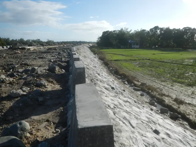 The image shows a concrete retaining wall in the middle of a dirt road, surrounded by stones,...