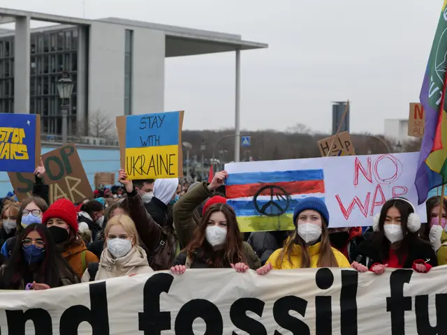 The image shows a group of people wearing masks and holding signs and flags in front of a building,...