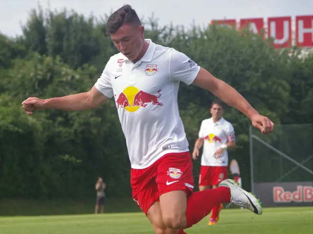 The image shows a man kicking a soccer ball on top of a grassy field, surrounded by trees and a...