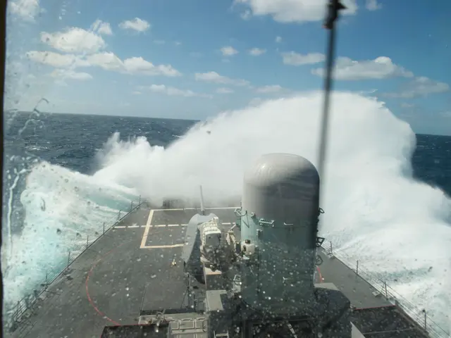 The image shows a ship in the ocean with a large wave crashing against it. On the deck of the ship,...