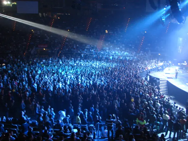 The image shows a large crowd of people in a concert hall, illuminated by bright lights and...