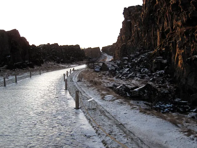 The image shows a snow-covered road winding through a rocky gorge, with a fence running alongside...