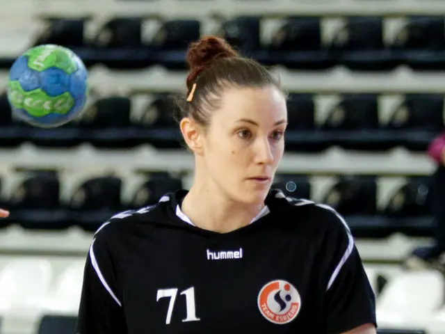 The image shows a woman standing on the floor, holding a soccer ball in her hands, with another...