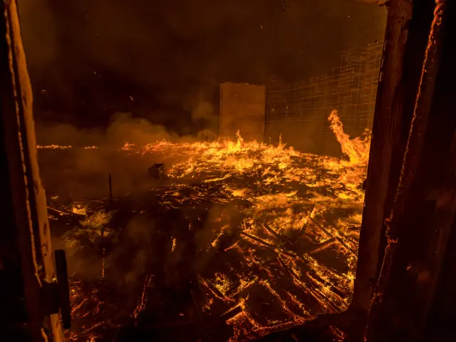The image shows a raging fire engulfing a building in the middle of a city at night, with smoke...