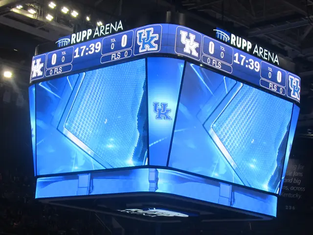 The image shows a large screen in the middle of a basketball court, surrounded by a group of people...