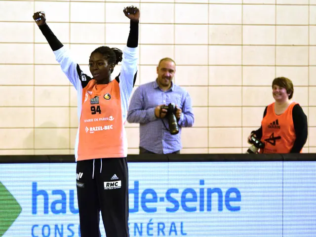 The image shows a woman standing on top of a basketball court with her arms in the air, surrounded...