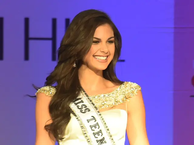 The image shows a woman in a white dress standing on a stage, smiling confidently. She is wearing a...