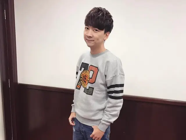 The image shows a young man standing on top of a red carpet, wearing a grey sweatshirt and blue...