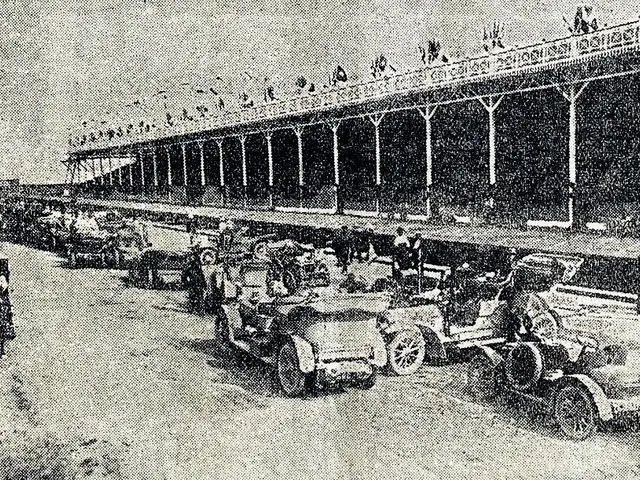 The image shows a black and white photo of a long line of cars parked in front of a building, with...