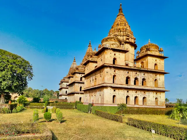 The image shows a large building in the middle of a lush green field, surrounded by trees, plants,...