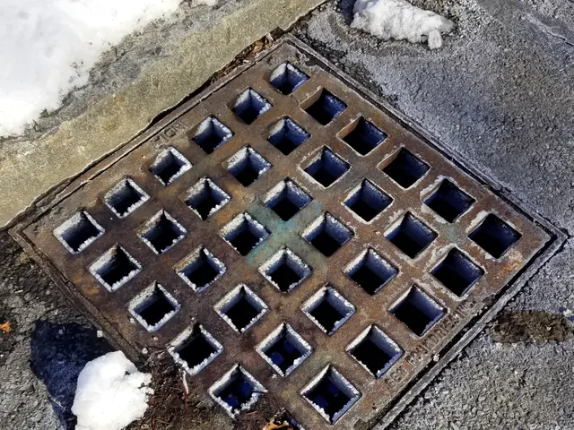 The image shows a metal manhole cover on the side of a road, with snow on the ground around it.