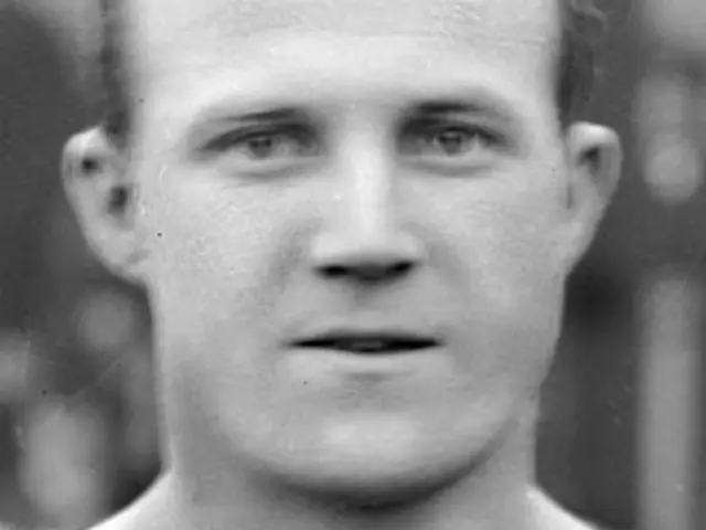 The image shows a black and white photo of a man wearing a t-shirt, with a blurred background. The...