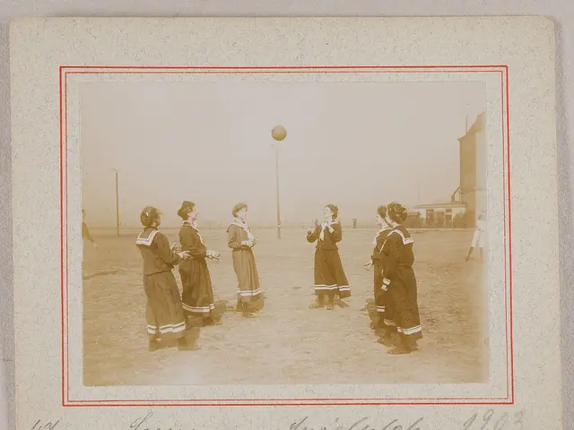 The image shows an old photo of a group of women playing soccer on the ground, with a ball in the...