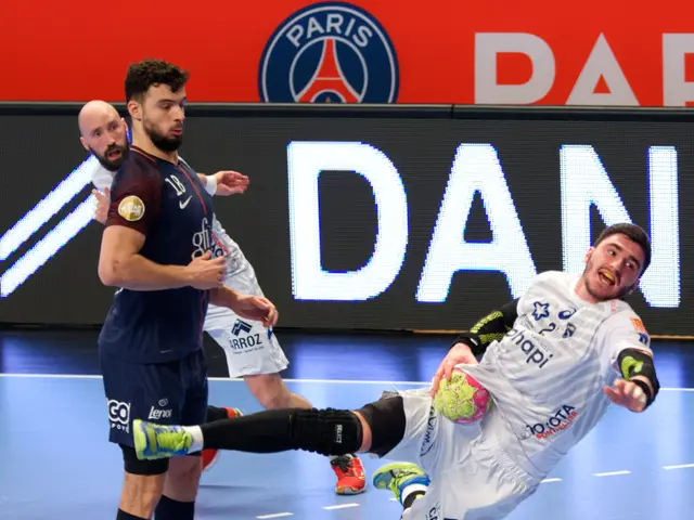 The image shows a group of men playing a game of handball on a court, with one of them holding a...
